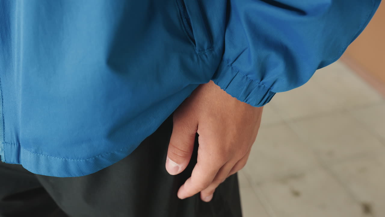 giacca blu, mano che esce dalla tasca, primo piano laterale piovoso che mostra il tessuto bagnato, dettaglio della cerniera e mano rilassata vicino ai pantaloncini, un movimento sottile cattura l'attesa prepartita, postura calma, manica in nylon strutturato