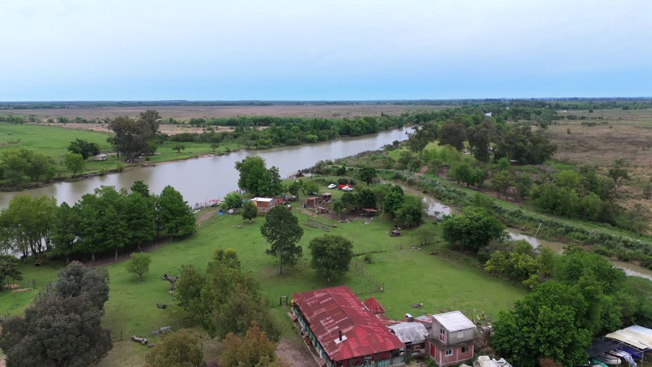 Aerial drone view of Martín Irigoyen Canal in the Buenos Aires Delta, Argentina, showing a rural riverside area with green pastures, trees, small houses, and calm brownish water under a cloudy sky