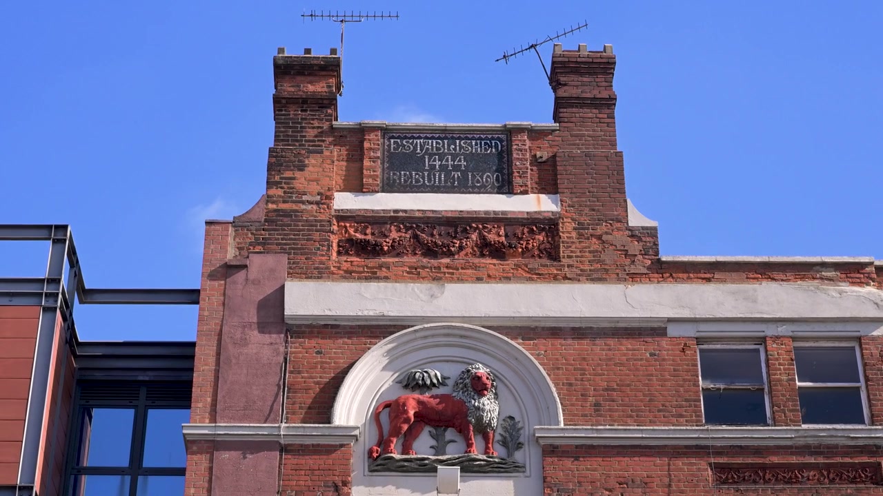 Il pub Red Lion su Kilburn High Road riempie l'inquadratura, con il suo stemma del leone rosso e la targa dello storico locale che si stagliano contro un cielo azzurro limpido