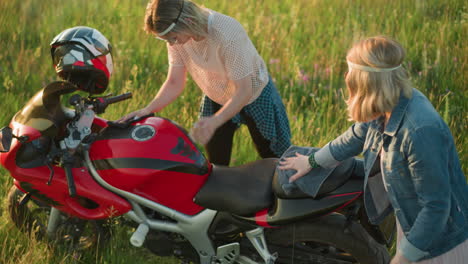 dos amigos son vistos limpiando una motocicleta roja en un campo abierto cubierto de hierba, uno se centra en limpiar el tanque de la bicicleta, mientras que la otra dama toca su cinta para la cabeza