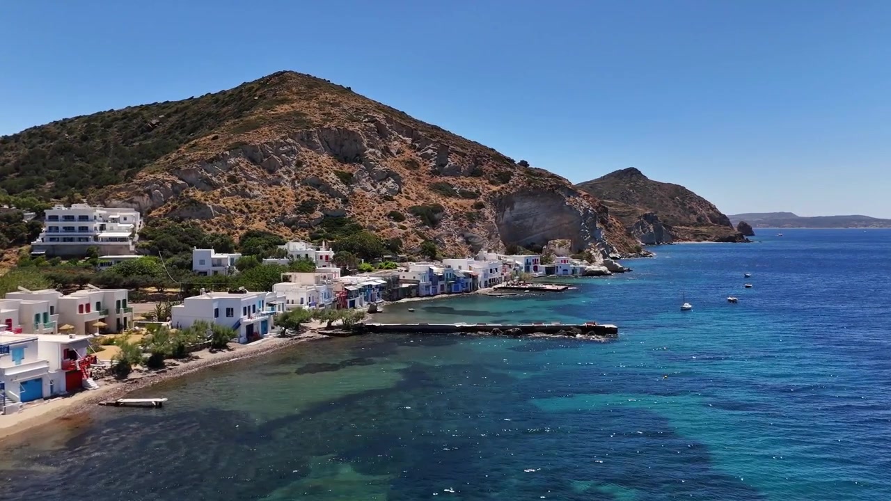 Il lungomare di Klima si estende accanto a un lungo molo in pietra mentre una vista aerea mostra case bianche e porte colorate sotto una collina rocciosa, con piccole barche a vela su acqua blu profondo sotto un sole limpido.