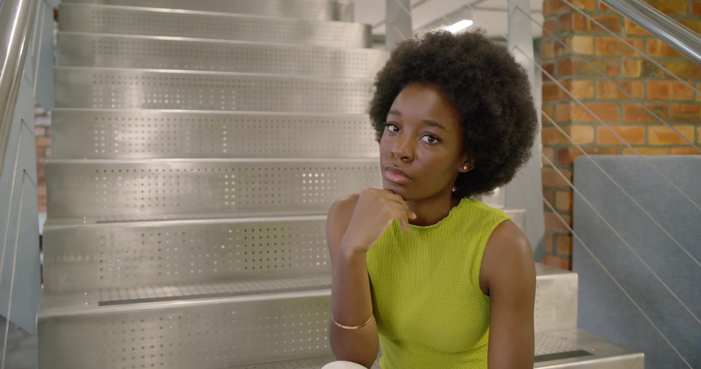 African American woman sitting on metal stairs in lime top, resting chin as camera tightens