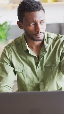 vertical video of african american man leaning on countertop in kitchen, using laptop