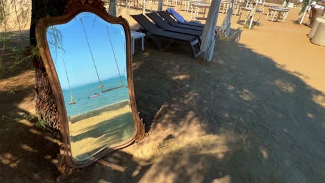 Imágenes-Inusuales-De-Un-Espejo-Apoyado-Contra-Un-árbol-En-La-Playa-Que-Refleja-Columpios-Y-Personas-Bañándose-En-El-Mar.