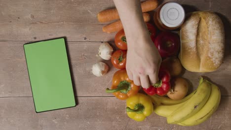 overhead studio shot of hand putting pepper in group of fresh food items with green screen digital tablet on wooden surface