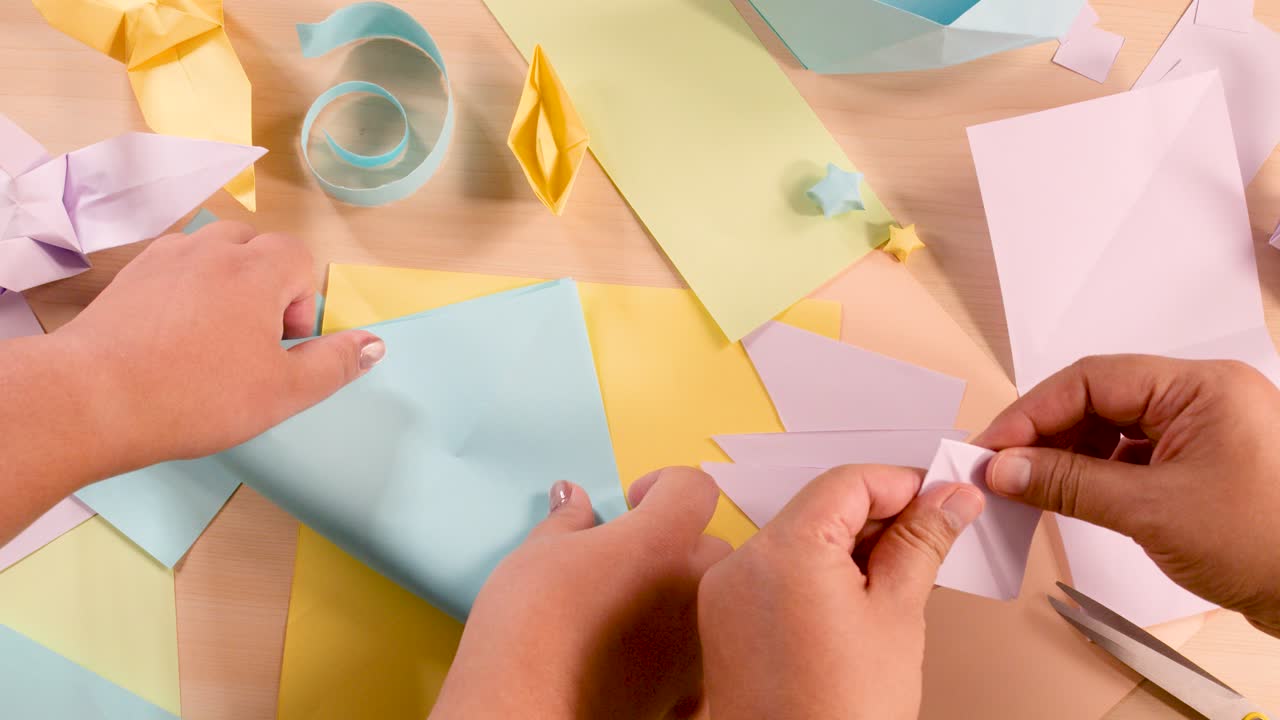 Two people fold pastel paper into origami shapes on a well-lit tabletop with craft supplies