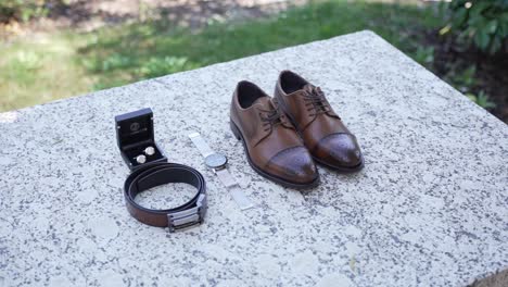 brown leather shoes, belt, watch, cufflinks placed on a stone table in an outdoor setting