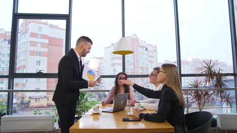small team of people working together sit at the table. male speaker in suit holds a paper with chart for collective group discussion. cityscape at backdrop.