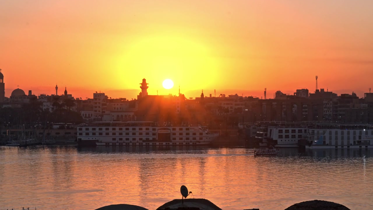 Scatto al tramonto su Luxor, Egitto, che mostra intensi raggi di sole che si irradiano nel cielo mentre il sole scende sotto l'orizzonte, proiettando un caldo bagliore arancione sul fiume Nilo e sulle navi da crociera ormeggiate