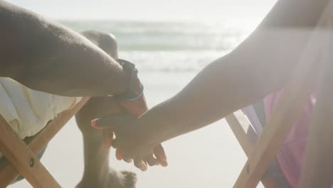 Senior-african-american-couple-sitting-on-deck-chairs,-holding-hands-at-beach,-in-slow-motion