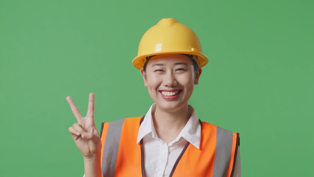 안전 헬을 입은 아시아 여성 엔지니어가 녹색 스크린 배경 스튜디오에 서 있는 동안 미소 짓고 평화의 몸짓을 보여줍니다.
