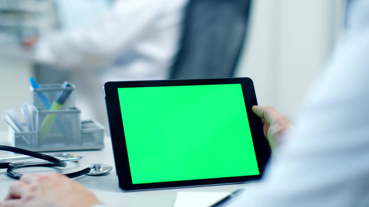 primer plano de un médico masculino que usa una tableta con pantalla verde, su asistente trabaja en el fondo. la oficina es brillante, blanca y moderna.