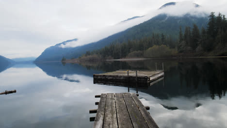 pov geht auf einem holzpier auf einer ruhigen seeoberfläche mit einer perfekten reflexion des himmels, der wolken, des berges und der bäume im wasser