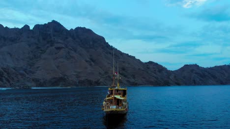 Un-Crucero-Anclado-Frente-A-La-Costa-De-La-Isla-Padar-Contra-Una-Montaña-Empinada-Y-Un-Cielo-Azul,-Cerca-De-Komodo-En-Indonesia,-Capturado-Durante-La-Noche