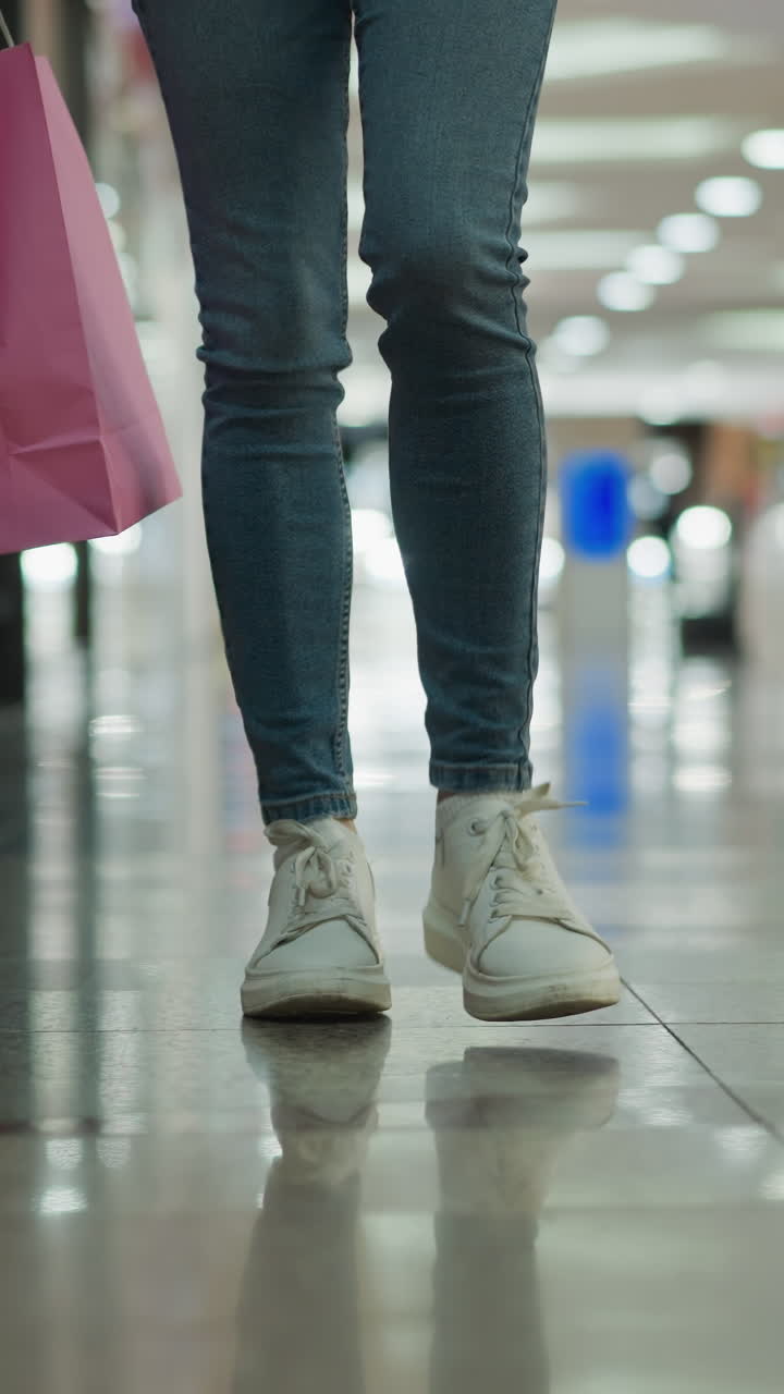 vista de la pierna de una mujer en vaqueros y zapatillas de lona que lleva bolsas de compras de menta y rosa en su mano derecha mientras camina a través de un centro comercial brillantemente iluminado con pisos de baldosas brillantes