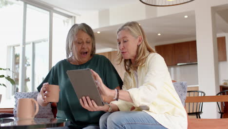 using tablet, two multiracial senior female friends sitting on couch and drinking coffee , at home