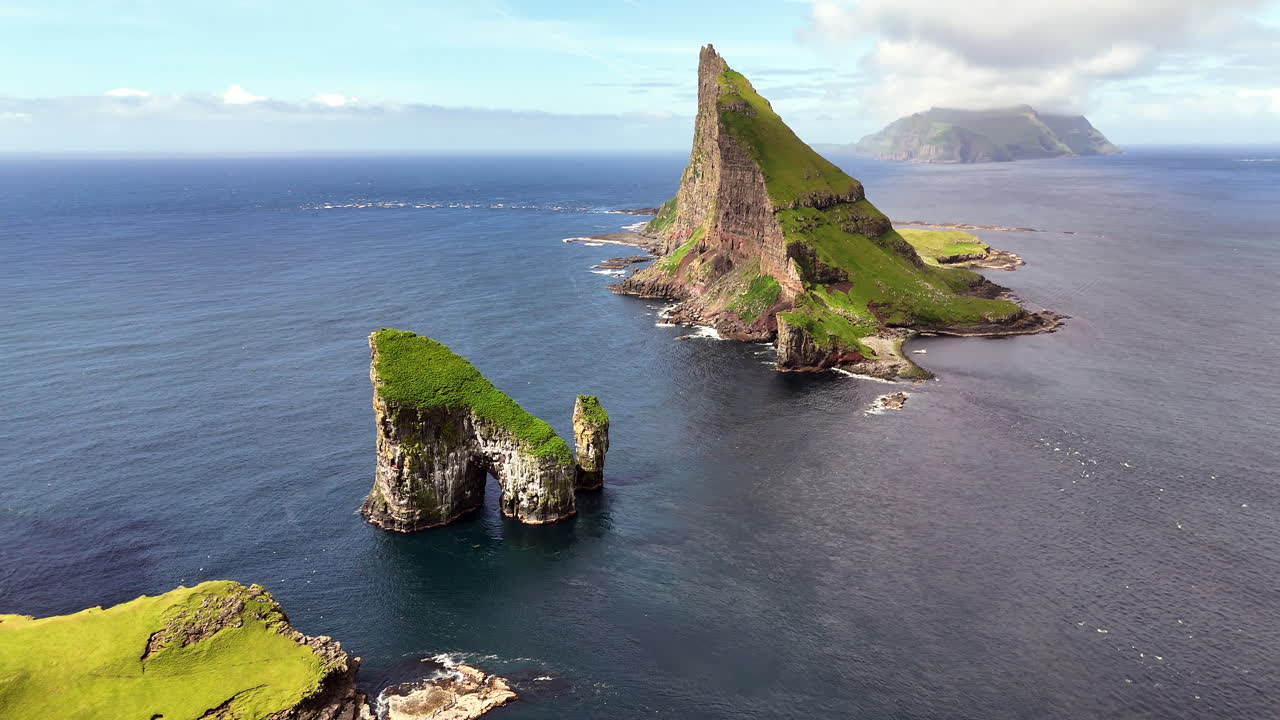 Cinematic aerial view of Drangarnir sea stacks rising dramatically from the Atlantic Ocean near Vágar, Faroe Islands, showcasing rugged cliffs, lush green slopes, and misty Nordic seascape