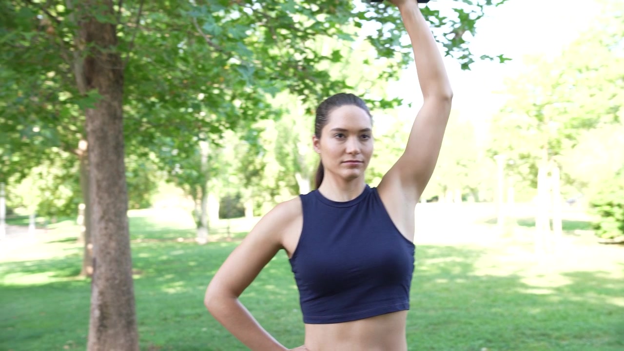 Una donna si allena con un manubrio in un parco, praticando esercizi di fitness all'aperto.