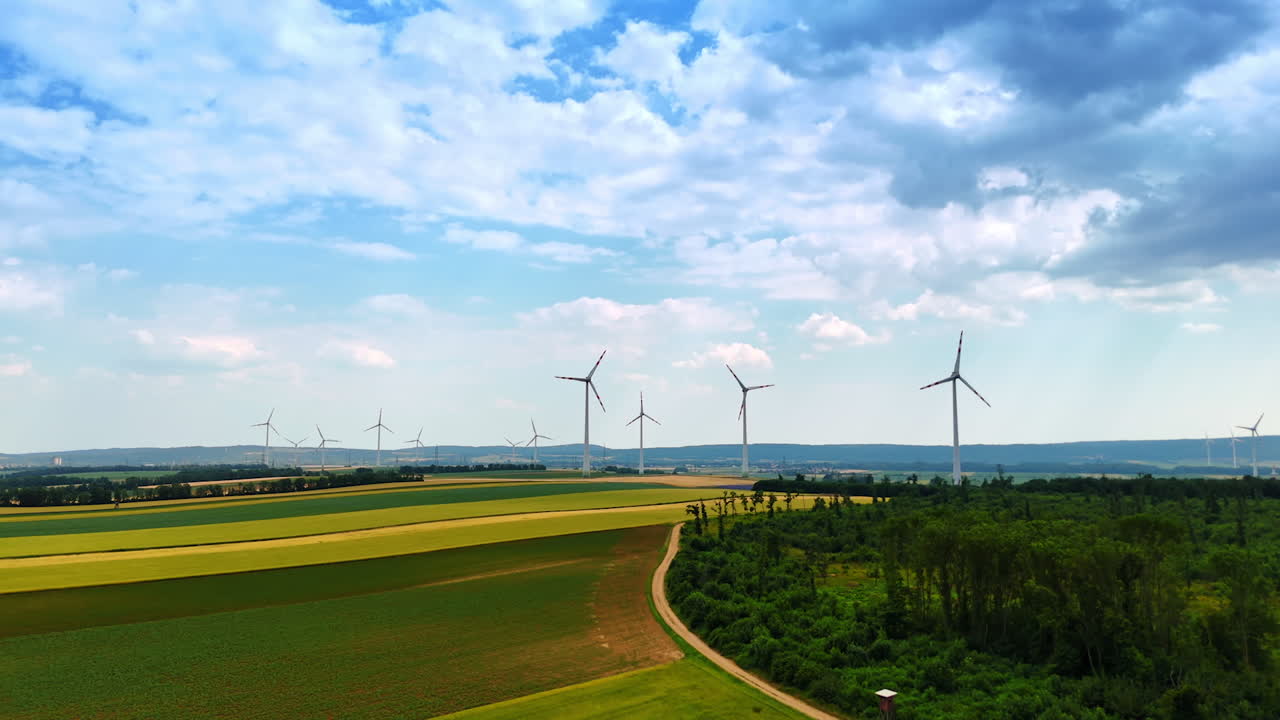 Beautiful countryside on summer day. Drone footage over the field and woods revealing view on the wind farms at backdrop. Clouds in the sky