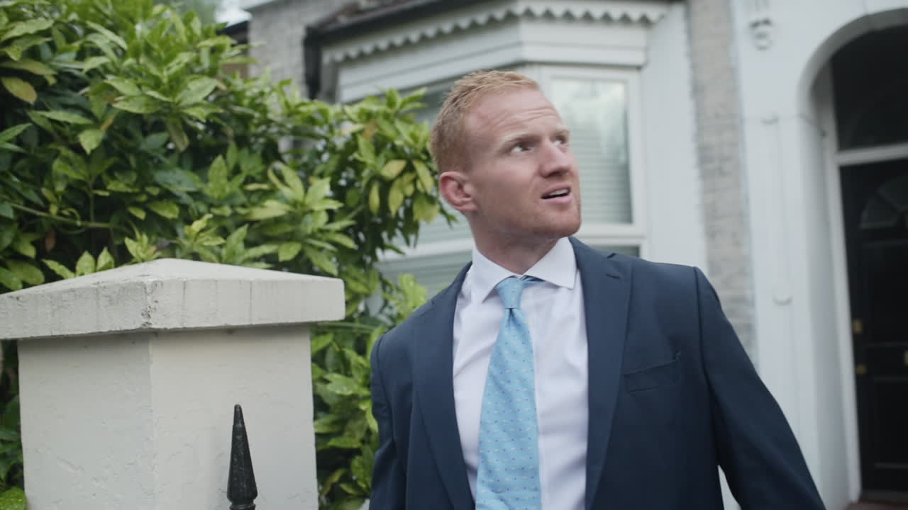 A middle-aged man puts on the jacket to his suit outside his home during his morning commute routine while walking from his front door to the street