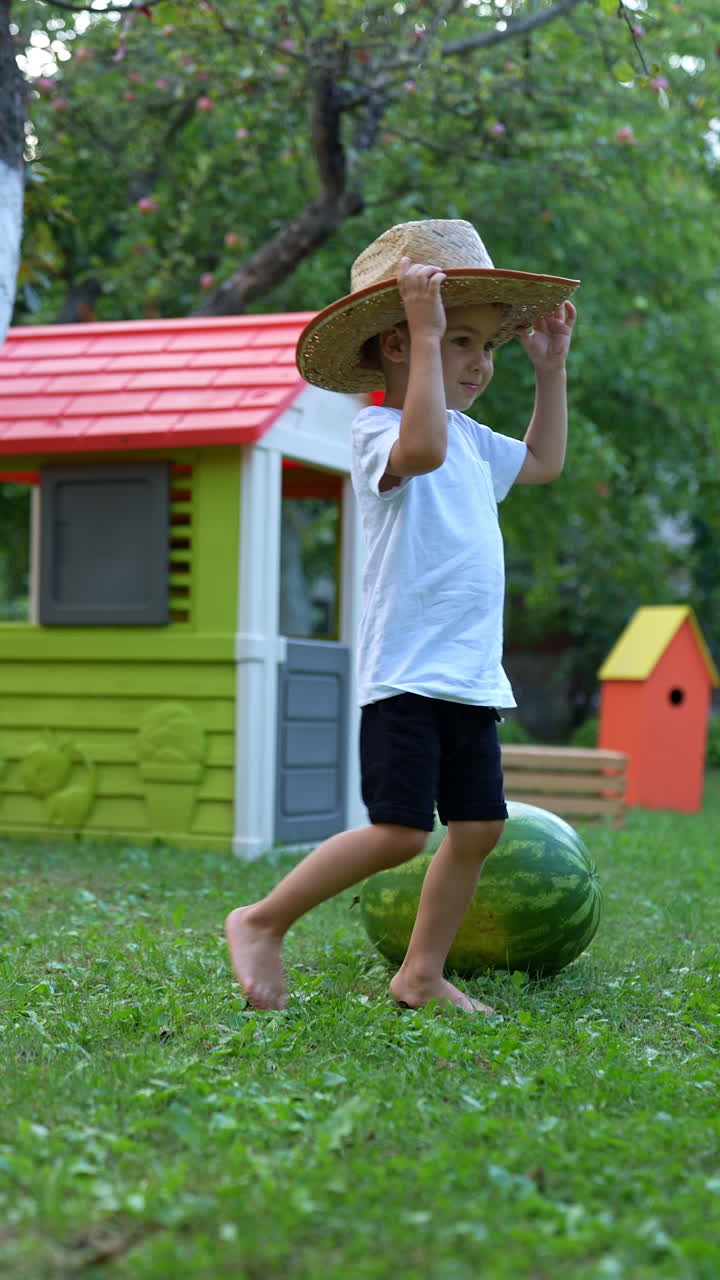 Little baby boy in big straw hat comes up to a huge watermelon in the garden. Kid puts his head to the berry. Vertical video.