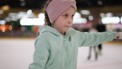 close-up of a lady in a mint green hoodie and pink headband, steering intently at the camera while ice skating, the background features blurred lights and people skating