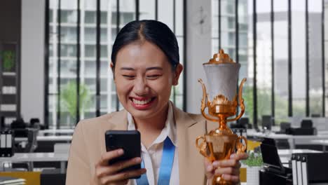 close up of asian business woman in a suit with a gold medal and trophy looking at smartphone then screaming goal celebrating in the office