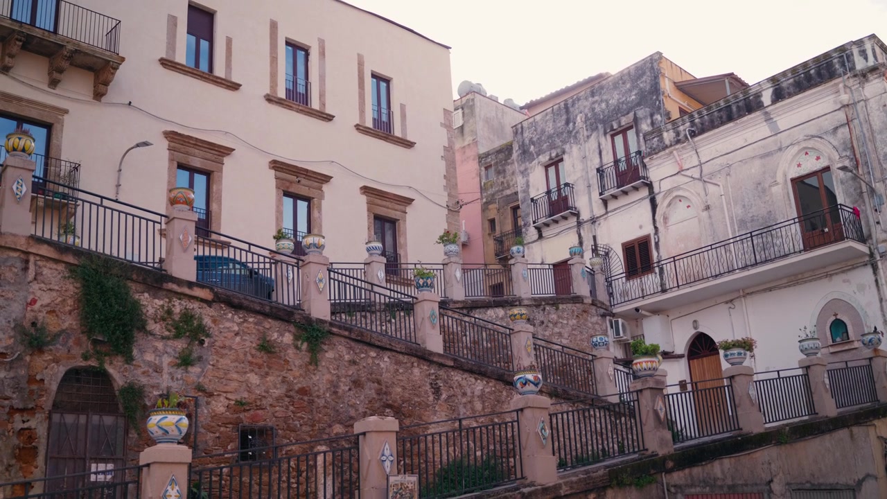 Una scalinata in pietra ornata, fiancheggiata da vasi in ceramica tradizionali e colorati, si snoda attraverso un cortile storico siciliano