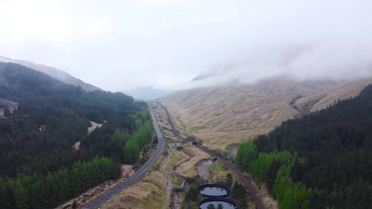 Aerial drone reversing or revealing footage flying over scottish brown grassy hills and pine forest mountains that have a road between them during a misy and foggy day. Pine trees are vibrant green.