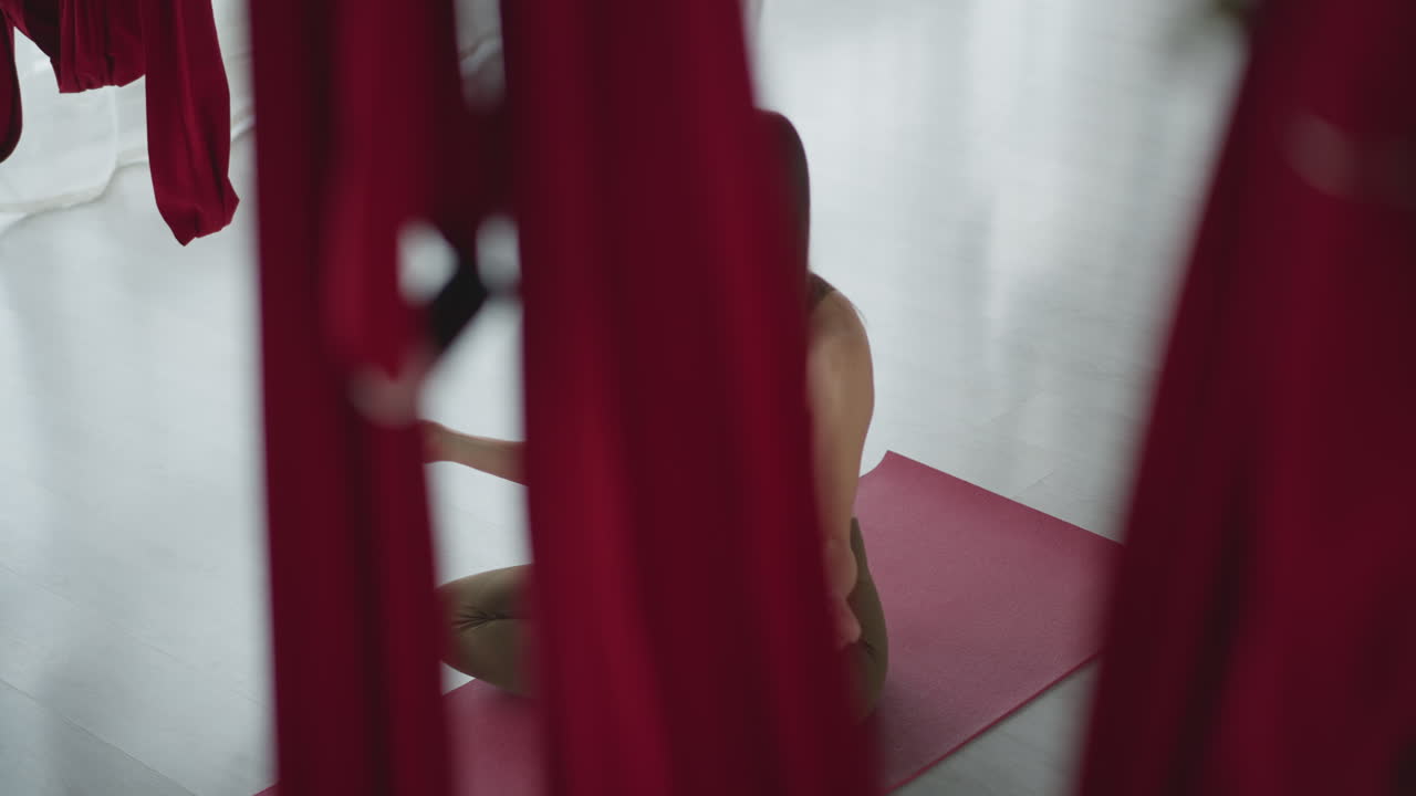 High angle shot of flexible woman in sports wear on yoga mat, legs crossed, placing hand on chest and stomach, practicing mindful breathing and meditation in studio with suspended silk hammocks