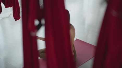 high angle shot of flexible woman in sports wear on yoga mat, legs crossed, placing hand on chest and stomach, practicing mindful breathing and meditation in studio with suspended silk hammocks