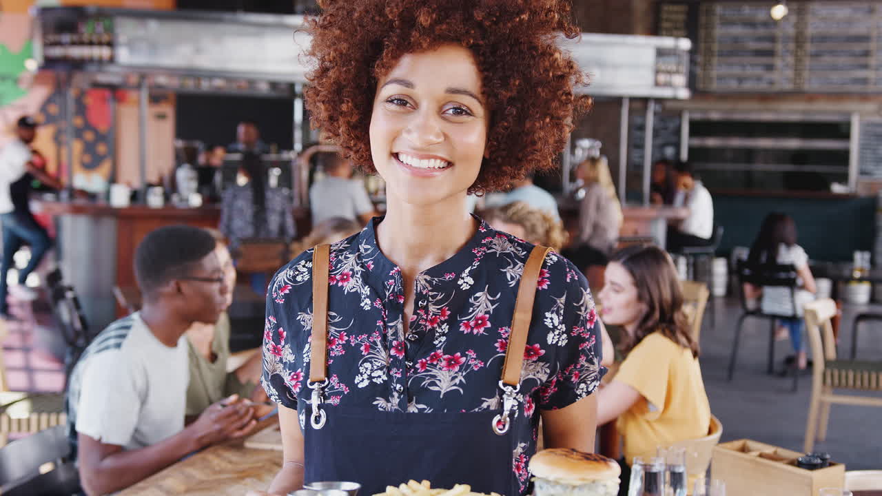 바쁜 술집 레스토랑에서 음식을 제공하는 웨이트리스의 초상화가 카메라에 미소 짓고 있습니다.