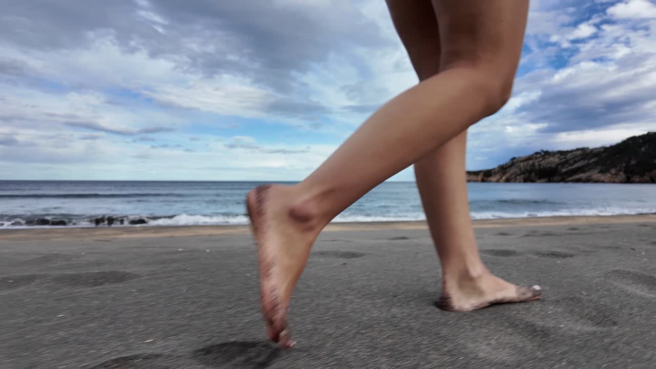 Slow-motion shot of bare legs walking along a sandy beach with waves and a cloudy sky behind