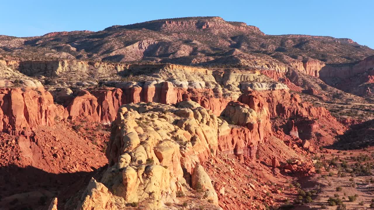 Una drammatica transizione con drone inizia dalle creste rocciose verticali di Abiquiu e panoramica per rivelare un orizzonte montuoso massiccio e distante. Questo vasto paesaggio cattura la vastità dell'aspro Sud-Ovest