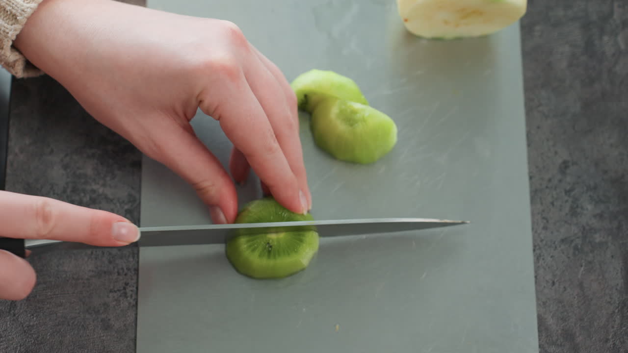 Schwangere Mutter bereitet Kiwischeiben auf einem Brett zu, Draufsicht auf Hände, die geschälte Früchte mit einem Wellenschliffmesser auf einer Matte schneiden, ordentliche Portionen angerichtet, helles Küchenlicht, achtsame Zubereitung eines pränatalen Snacks