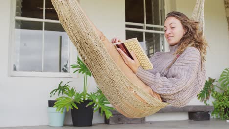 smiling caucasian woman lying in hammock reading book on terrace