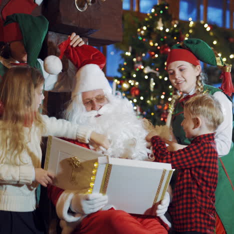 children and elves touch santa's white beard and red cap on christmas background. santa claus is sitting with album and having fun with two little kids and fairy elves indoors.