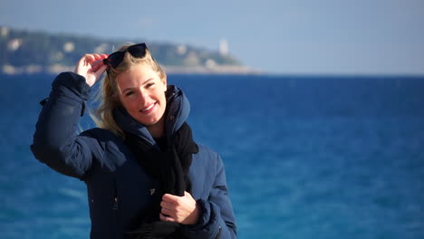 Close-up-of-a-young-blonde-woman-laughing-to-the-camera,-the-blue-sea-in-soft-focus-behind-her