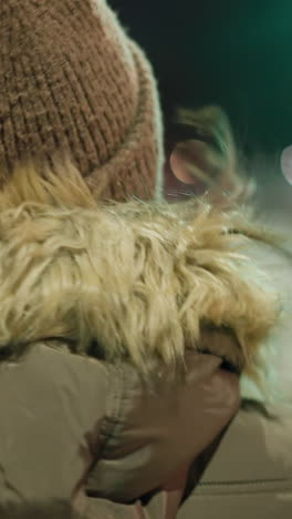 first-person view of a woman in a winter coat with a fur hood, facing a green traffic signal at a nighttime urban intersection with blurry lights and snowy background. walking slowing across the road