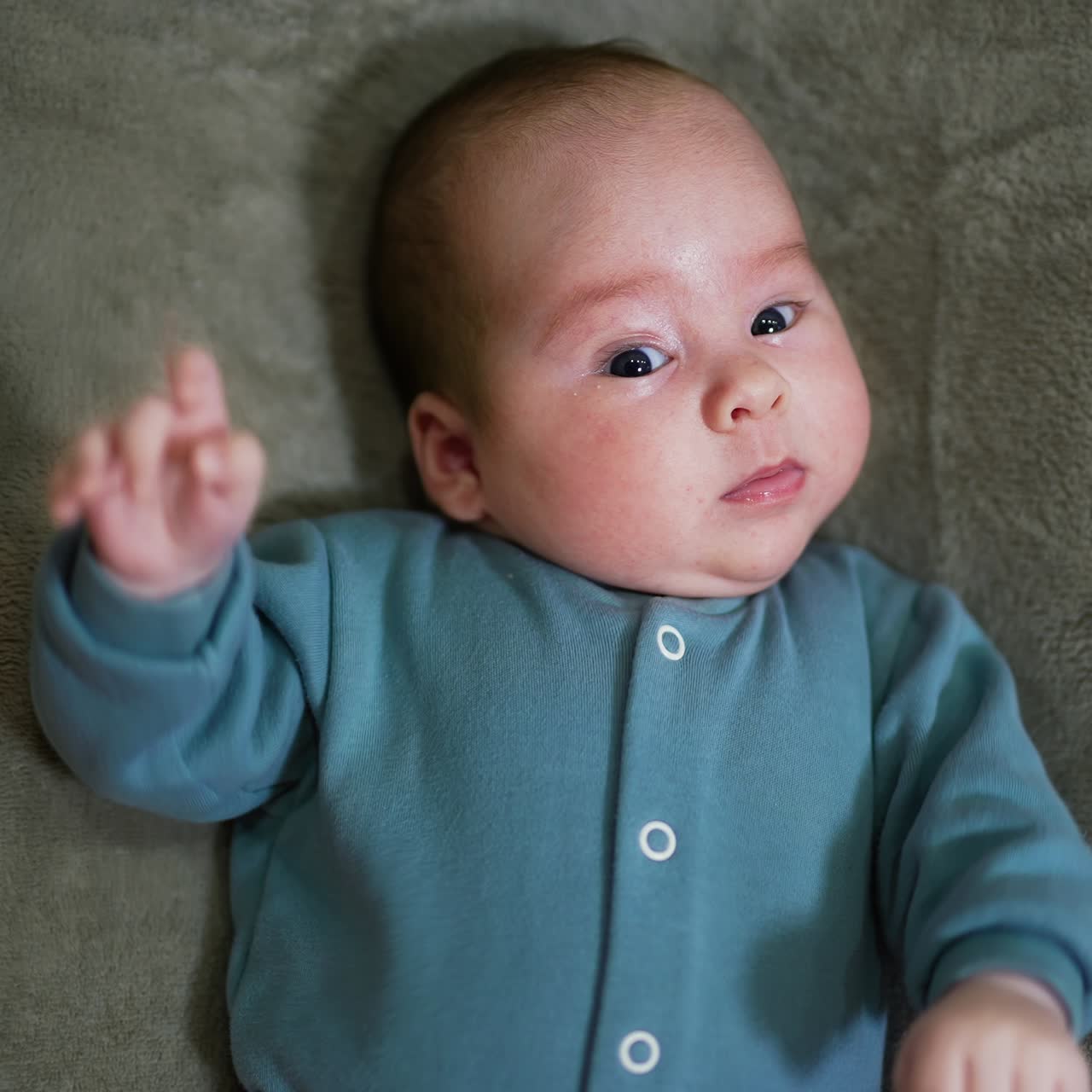 Bayi menggemaskan menatap kamera dengan curiga. Bayi laki-laki manis berpakaian biru berbaring di tempat tidur. Close up. Latar belakang abu-abu