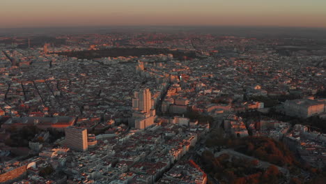 Aerial-panoramic-view-of-city-at-twilight-from-height.-Buildings-lit-by-orange-sunset-light.