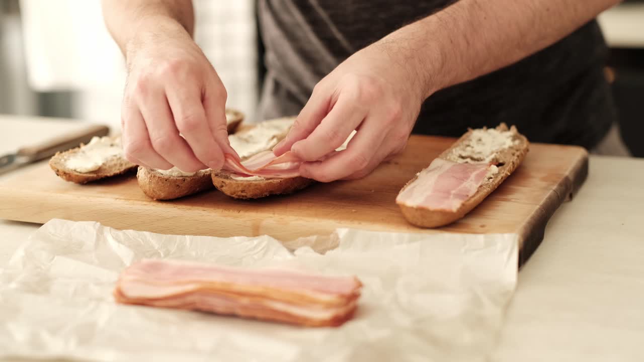 Caucasian male carefully placing bacon strips on sliced baguette in the kitchen
