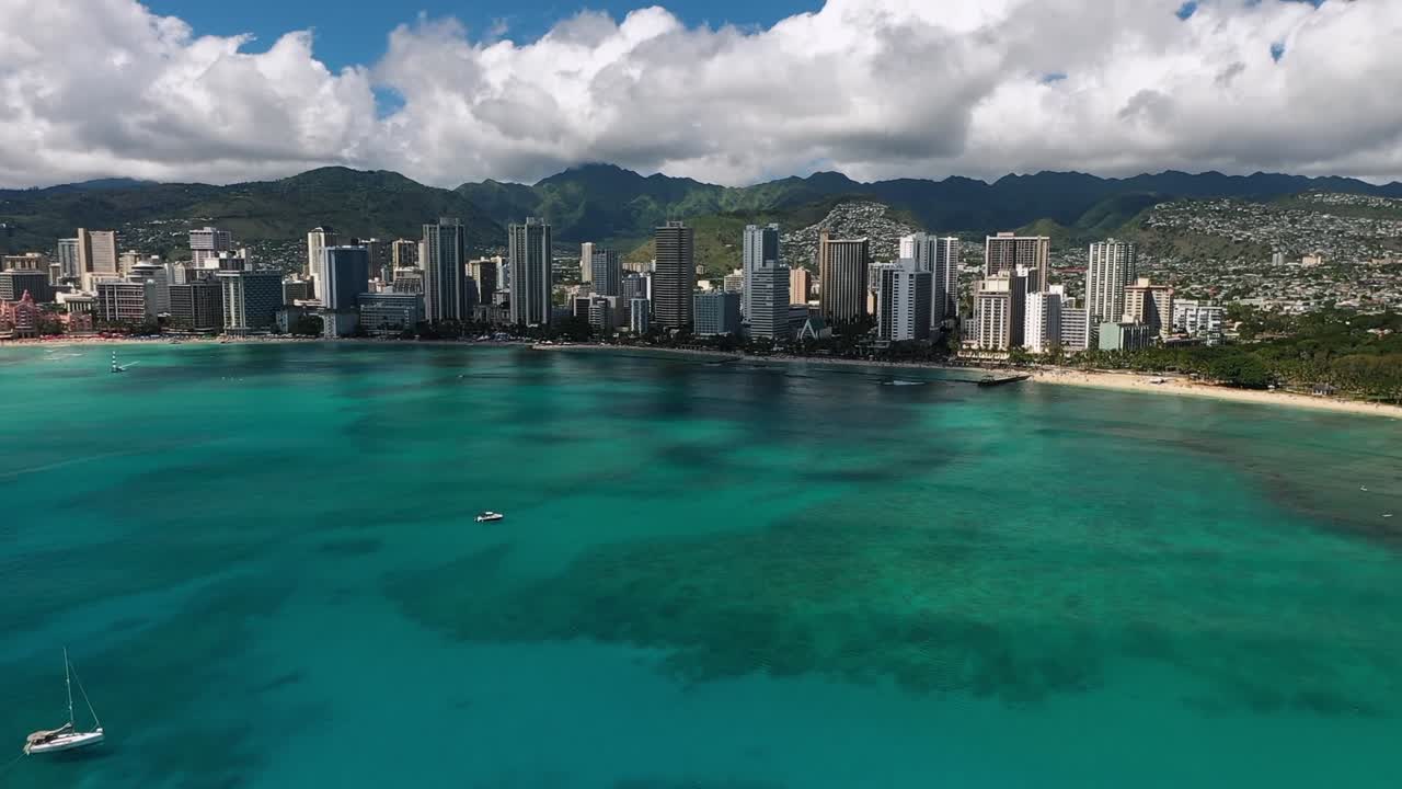 Video aereo da drone che mostra lo skyline di Honolulu con acqua turchese dell'oceano, costa sabbiosa e barche a vela vicino a Waikiki Beach sull'isola di Oahu sotto un cielo soleggiato e luminoso alle Hawaii