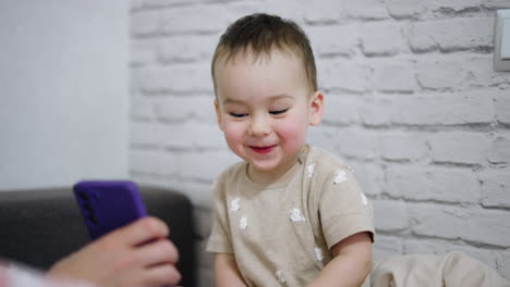 adorable little boy is amused by something shown on the phone. cute baby smiles and giggles sweetly. white wall backdrop.