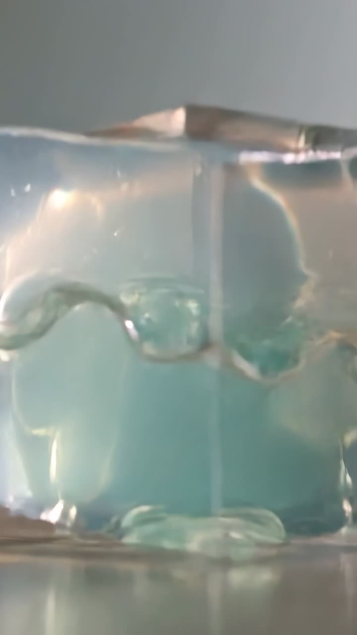 Video verticale: Un cubo di gel turchese increspato si adagia su un piatto lucido, rivelando bolle interne.