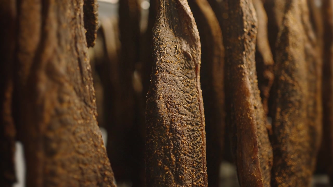 Lunghe strisce di biltong essiccato pendono fitte, le loro ruvide texture di crosta speziata riempiono l'inquadratura in una luce calda.