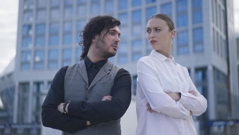 argued business couple standing back to back on city street looking at each other turning away in slow motion. blurred downtown building at background. conflict and problem concept.