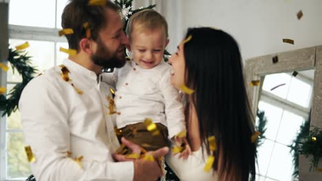 Young-mother,-father-and-their-little-son-celebrating-with-shooting-confetti-on-Christmas-Eve-at-home,-kissing-their-cute-child