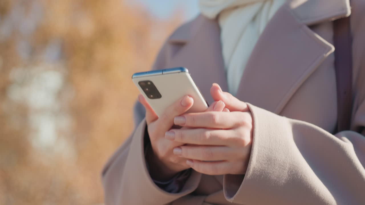 Hände einer weißen Frau halten Smartphone in Nahaufnahme, gepflegte Nägel tippen auf den Bildschirm, Ärmelbund eines Wollmantels sichtbar, warmes Herbst-Bokeh, gezielte Gesten und das Einstecken des Telefons, dezente Signale für Privatsphäre und Sicherheit.