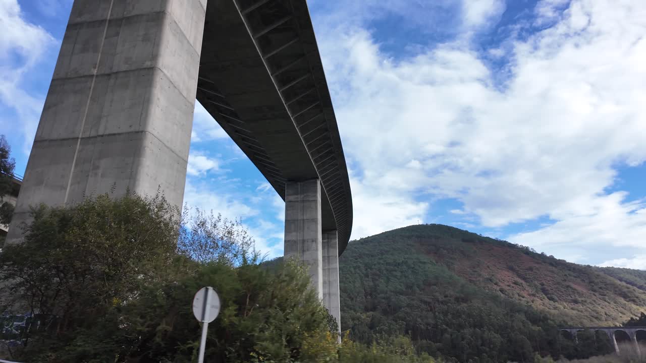 Pilastri enormi di un ponte in cemento si ergono tra gli alberi sotto un cielo nuvoloso, evidenziando la scala e il contrasto ambientale